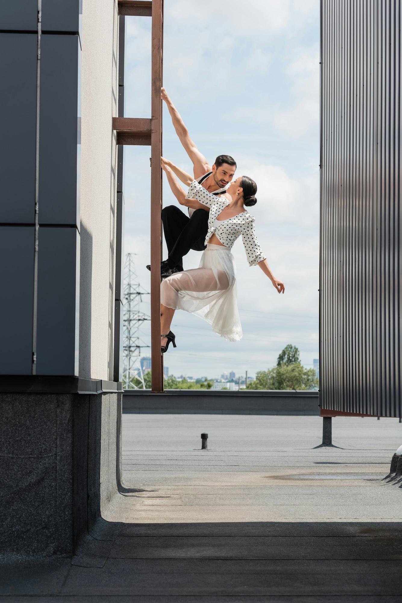 Professional ballroom dancers looking at each other while posing on ladder on rooftop of building